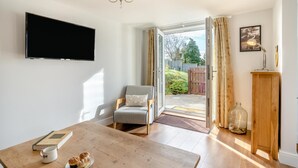 Una sala de estar acogedora con un televisor de pantalla plana, una mesa de centro de madera con un libro y pasteles, un sillón cómodo y vistas a un jardín a través de una puerta de cristal.