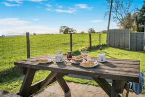 Un conjunto de mesa y sillas de picnic de madera al aire libre, con platos de comida, vasos y un tarro de mermelada.