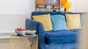 Ein blaues Sofa mit gelben und blauen Kissen, ein runder Beistelltisch mit goldener Rahmen, auf dem sich eine Schale mit Obst und Gebäck befindet, und eine gelbe Lampe im Hintergrund.