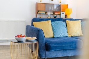 A blue sofa with yellow and blue cushions, a round gold-framed side table with a bowl of fruit and pastries, and a yellow lamp in the background. - 1 Bed in Cromer (oc-1844) (Cromer)