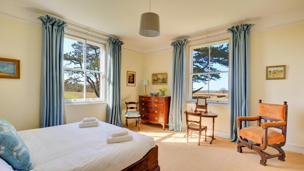 A bedroom with a large bed, a wooden dresser, a chair, and a desk with a lamp. There are two windows with curtains, and a painting on the wall. - 6 Bed in Lower Bodham (oc-1830) (Bodham)