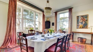 A dining room with a large table set for six, surrounded by chairs, with a patterned carpet, large windows, and a chandelier. - 6 Bed in Lower Bodham (oc-1830) (Bodham)