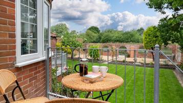 A balcony with wicker furniture, a table, and a view of a well-maintained garden and brick wall.