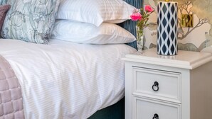 Una cama con ropa de cama blanca y cabecera azul, una mesita de noche con lámpara, y una pared con estampado de árbol.
