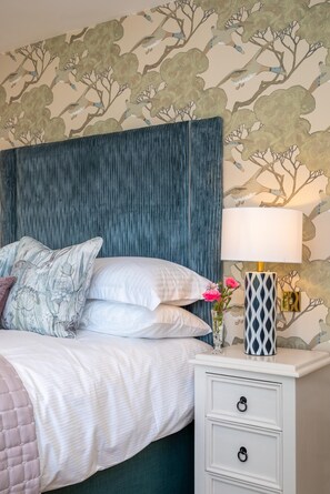 A bed with white bedding and a blue headboard, a bedside table with a lamp, and a wall with a tree pattern. - 2 Bed in Beccles (oc-rilo) (Beccles)
