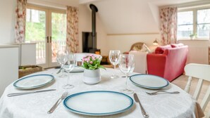 Mesa de jantar posta com pratos, talheres e copos de vinho, rodeada por uma sala de estar aconchegante com sofá e lareira.