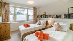 Quarto com duas camas, uma cômoda de madeira, uma mesa de cabeceira com luminária e uma estante de livros.