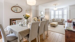 A dining area with a table set for four, surrounded by wicker chairs, a vase with flowers, and a bookshelf. - 4 Bed in Southwold (oc-23str) (Southwold)