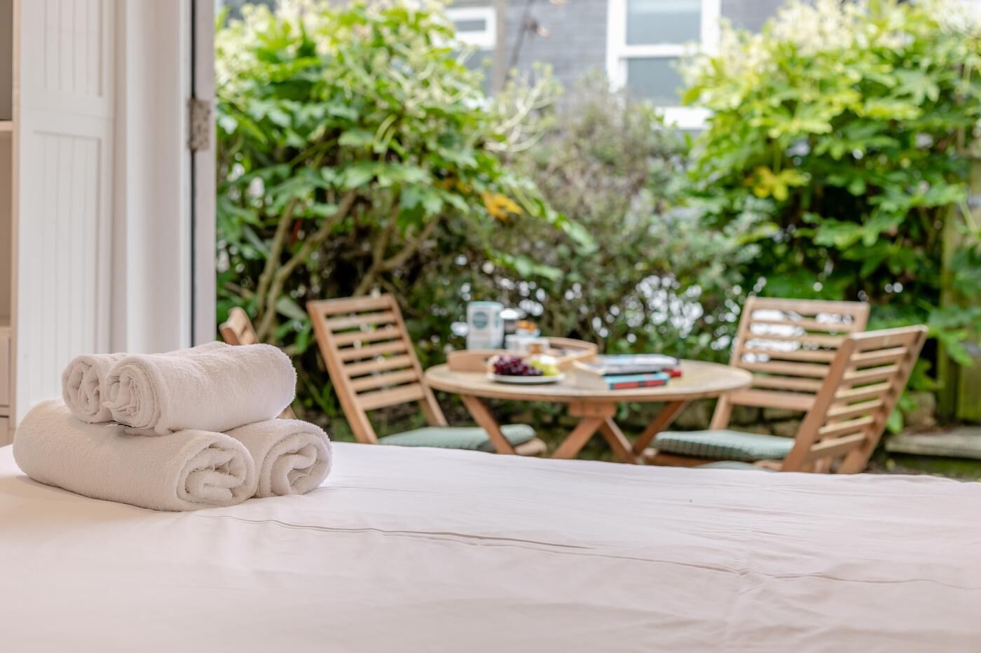 A neatly made bed with rolled white towels, a round wooden table with chairs, and a view of lush greenery outside.