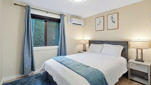 Een slaapkamer met een bed, twee nachtkastjes, een raam met gordijnen, een airconditioning en kunst aan de muur.
