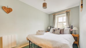 Un letto rifatto con copriletto bianco, un comodino con una lampada, una finestra con tende e una decorazione a forma di cuore sul muro.
