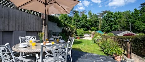 An outdoor patio with a table and chairs, a large umbrella, and a garden in the background.
