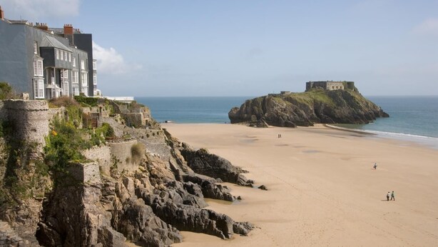 Une plage côtière avec une fortification en pierre, un grand bâtiment doté de nombreuses fenêtres et une structure ressemblant à un château, située sur un promontoire rocheux.