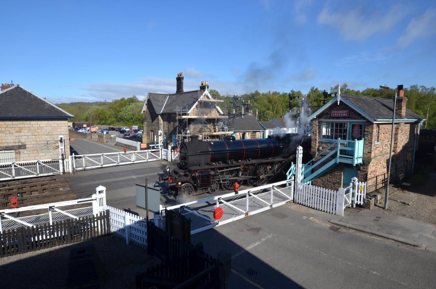 A steam locomotive at a railway crossing with a station building and a pedestrian bridge.