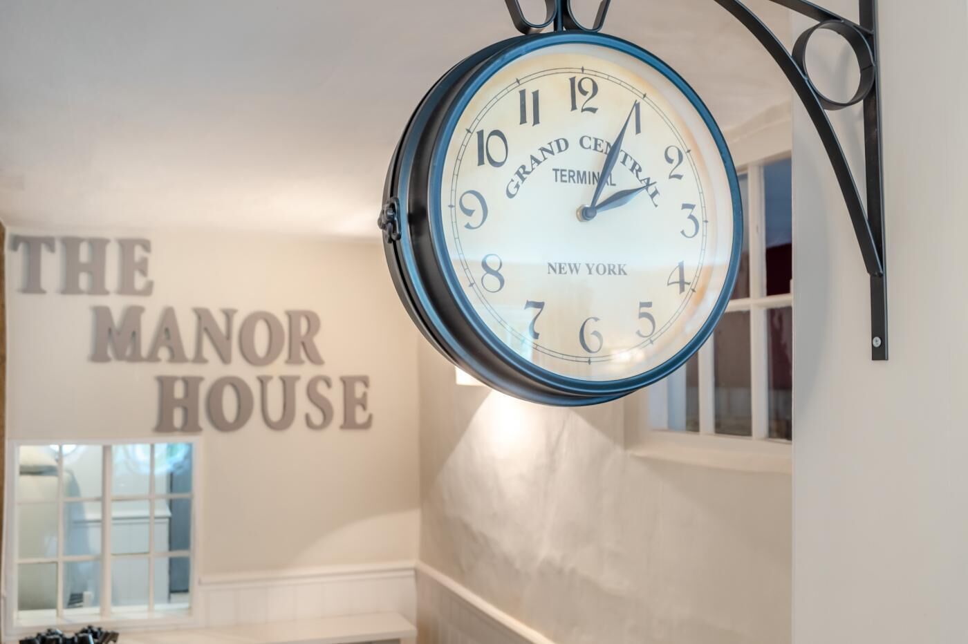 A clock with "Grand Central Terminal New York" on its face, mounted on a wall with a sign reading "The Manor House" in the background.
