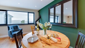 A dining area with a round wooden table set for two, featuring a bottle of wine, a glass, a plate, a baguette, cheese, and a loaf of bread. There is a vase with flowers and a chair in the background. - 5 Bed in Woolacombe (oc-alba12) (Woolacombe)