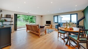 A spacious living area with a wooden sofa, a dining table with chairs, a television, and a large window offering a view of greenery. - 5 Bed in Woolacombe (oc-alba12) (Woolacombe)
