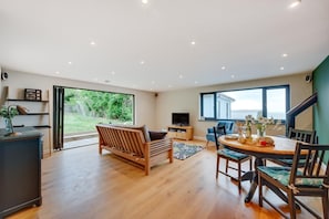 Amplia sala de estar con un sofá de madera, una mesa de comedor con sillas, un televisor y un gran ventanal con vistas a la vegetación.
