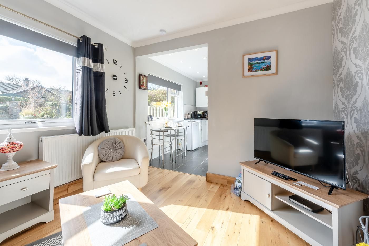 A modern living room with a flat-screen TV, a sofa, a dining area, and a kitchen in the background.