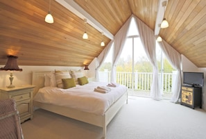 A bedroom with a wooden ceiling, a bed with white bedding, a nightstand, a lamp, a television, and a balcony with curtains.