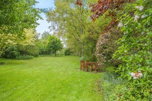 A grassy outdoor area with a wooden table and chairs, surrounded by dense foliage and trees. - 3 Bed in Ufford (oc-sfmea) (Ufford)
