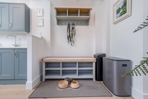 Een kleine keuken met grijze kastjes, een ingebouwde bank met opbergruimte, een prullenbak en voerbakken voor huisdieren op een vloerkleed.