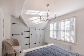 Een slaapkamer met een schuine kap, een kroonluchter, een bed, een stoel, een kledingkast en een raam met jaloezieën.
