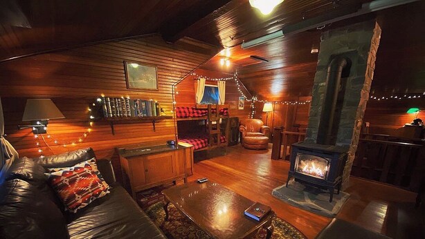 A cozy cabin interior with a fireplace, leather sofa, wooden walls, and a bookshelf.