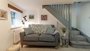 Een gezellige woonkamer met een grijze bank, een klein bijzettafeltje met een vaas, een trap en ingelijste foto's aan de muur.