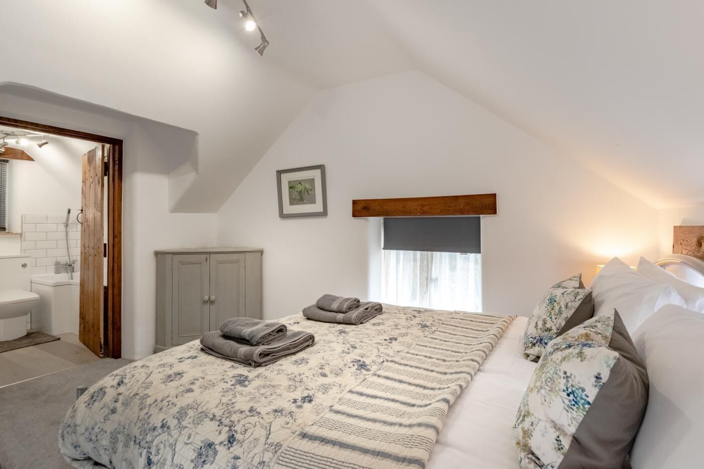 A bedroom with a large bed, a wooden headboard, a window with curtains, a wardrobe, and a bathroom visible through an open door.