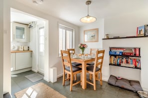 A dining area with a table set for four, a bookshelf with books, and a kitchen visible in the background. - 2 Bed in Orford (oc-sfcto) (Orford)