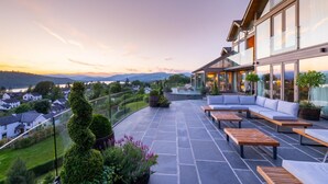 Un bâtiment moderne doté d’une façade en verre, d’un espace extérieur aménagé pour s’asseoir et offrant une vue sur la ville et les collines.