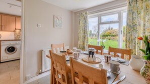 A dining area with a table set for four, wooden chairs, a view of a garden through large windows, and a kitchen area with a washing machine. - 3 Bed in Frostenden (oc-sfhms) (Frostenden)