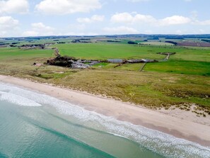 Luftaufnahme einer Küstenregion mit Sandstrand, grünen Feldern und einer Anlage mit mehreren Gebäuden.
