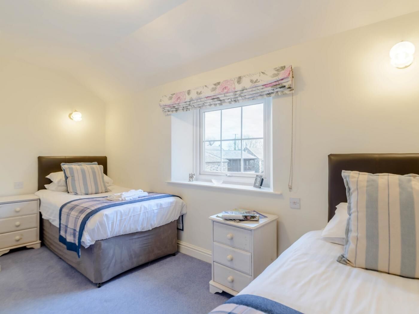 Une chambre avec deux lits, une fenêtre agrémentée d’une lambrequin à motifs floraux, une table de chevet avec un livre, et une commode avec des tiroirs.