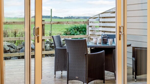 A glass door leading to an outdoor patio with wicker furniture and a coffee table, overlooking a grassy field. - 2 Bed in Alnwick (oc-80494) (Newton-by-the-Sea)