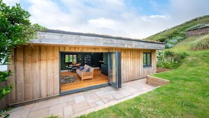 Una casa moderna con facciata in legno, un patio con divano e tavolo, e un giardino fiorito.