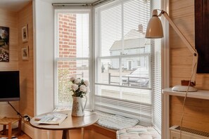 A cozy room with a large window, a small table with a vase of flowers, a lamp, and a television. - 4 Bed in Aldeburgh (oc-sfcrr) (Aldeburgh)