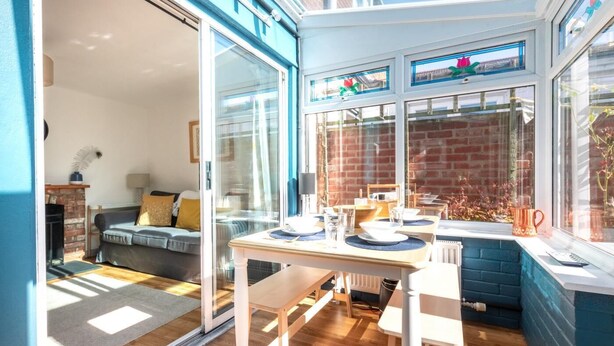 A bright, sunlit room with a dining table set for two, a blue-framed window, and a brick wall. - 2 Bed in Woodbridge (oc-48sc) (Woodbridge)