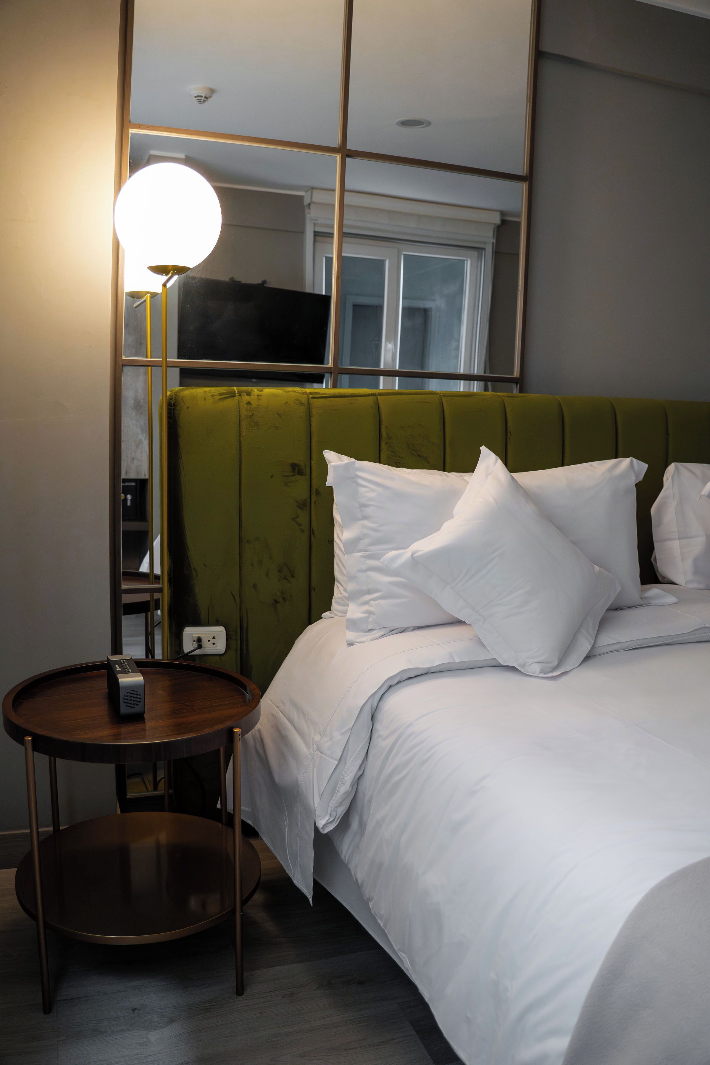 Hotelkamer met een groen bekleden hoofdeinde, witte beddengoed, een nachtkastje met een lamp en een stopcontact.