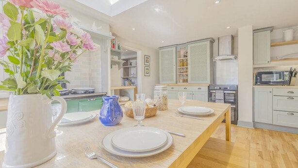 A modern kitchen with a wooden table set for dining, a white vase with flowers, and a blue pitcher. - 3 Bed in Woodbridge (oc-sfdor) (Woodbridge)