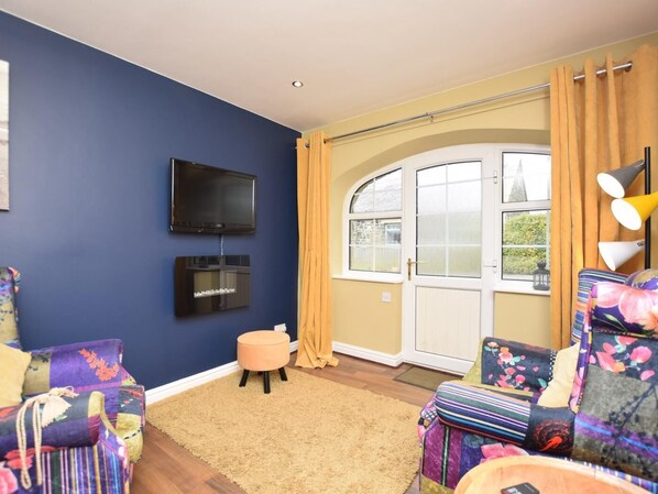 A living room with a blue wall, a flat-screen TV, a sofa with floral cushions, a round ottoman, and a window with yellow curtains. - 1 Bed in Alnwick (oc-80486) (Newton-by-the-Sea)