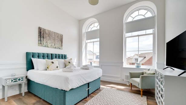 Une chambre moderne avec un lit recouvert d’un tissu turquoise, du linge de lit blanc et une grande fenêtre donnant sur la ville.
