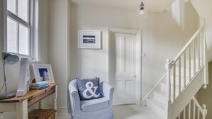 A hallway with a chair, a desk with a lamp, framed pictures, and a staircase. - 4 Bed in Aldeburgh (oc-sfhan7) (Aldeburgh)
