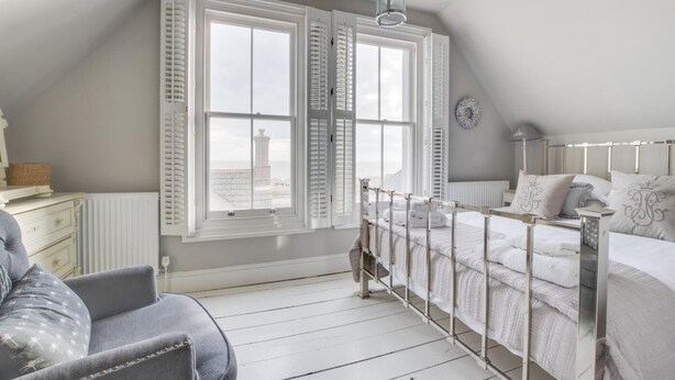 A bedroom with a bed, a chair, a window with shutters, a radiator, and a view of a tower. - 4 Bed in Aldeburgh (oc-sfhan7) (Aldeburgh)