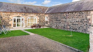 A stone building with a grassy courtyard, a table and chairs, and a gravel path. - 4 Bed in Alnwick (oc-80496) (Newton-by-the-Sea)