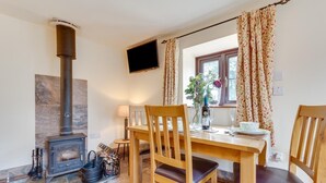 Een gezellige kamer met een houtkachel, een eettafel met stoelen, een aan de muur bevestigde televisie en een raam met gordijnen.