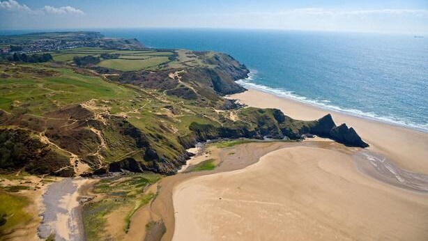 Luchtbeeld van een kustlandschap met een zandstrand, groene velden en een ruige kustlijn.