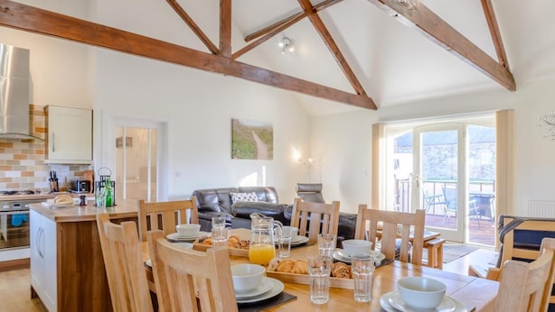 A spacious kitchen with wooden beams, a dining area with wooden chairs, a black leather sofa, and a view of the outdoors through large windows. - 3 Bed in Alnwick (oc-80489) (Newton-by-the-Sea)