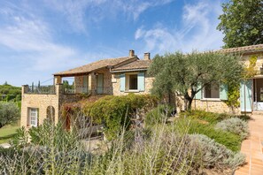 Una casa in pietra con un balcone, immersa in un giardino ricco di piante e un cielo azzurro intenso.